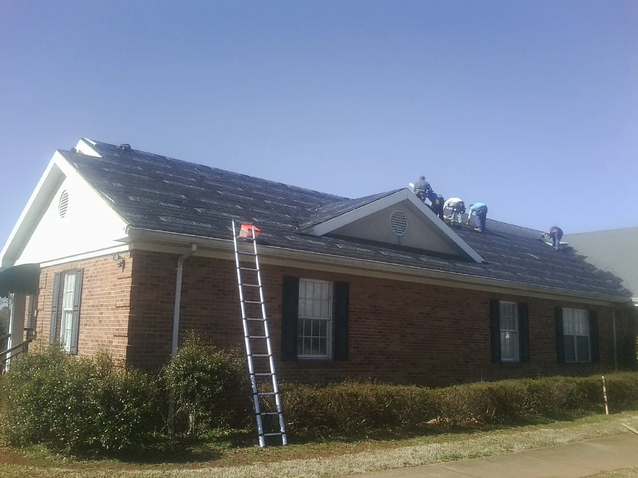 Crew tearing shingles off a brick ranch roof with a ladder set — Metal Roof Repair work by our crew in Clarence