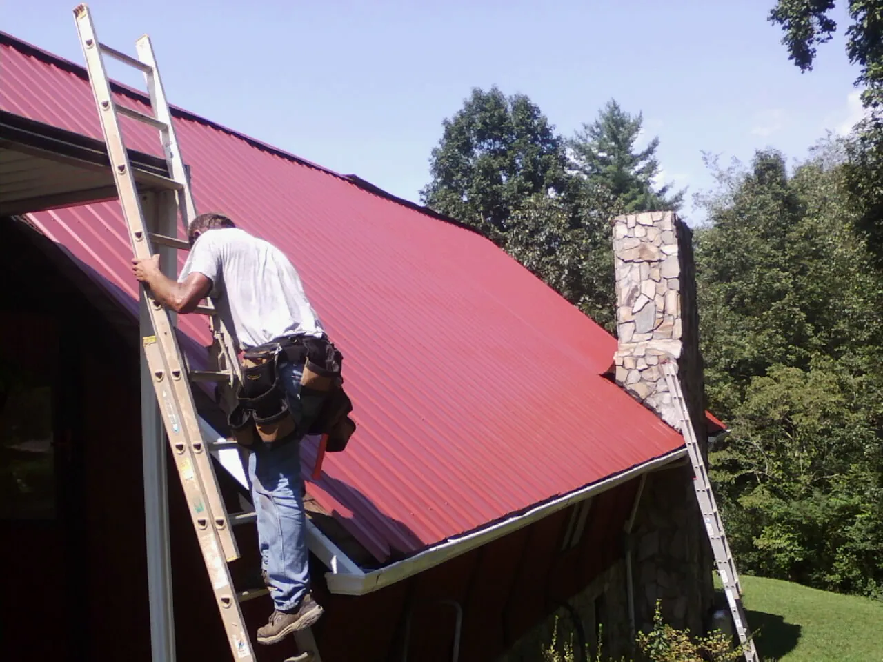 Roofer on a ladder beside a red metal roof and stone chimney — Soffit & Fascia Repair by Roofing Brothers in Clarence